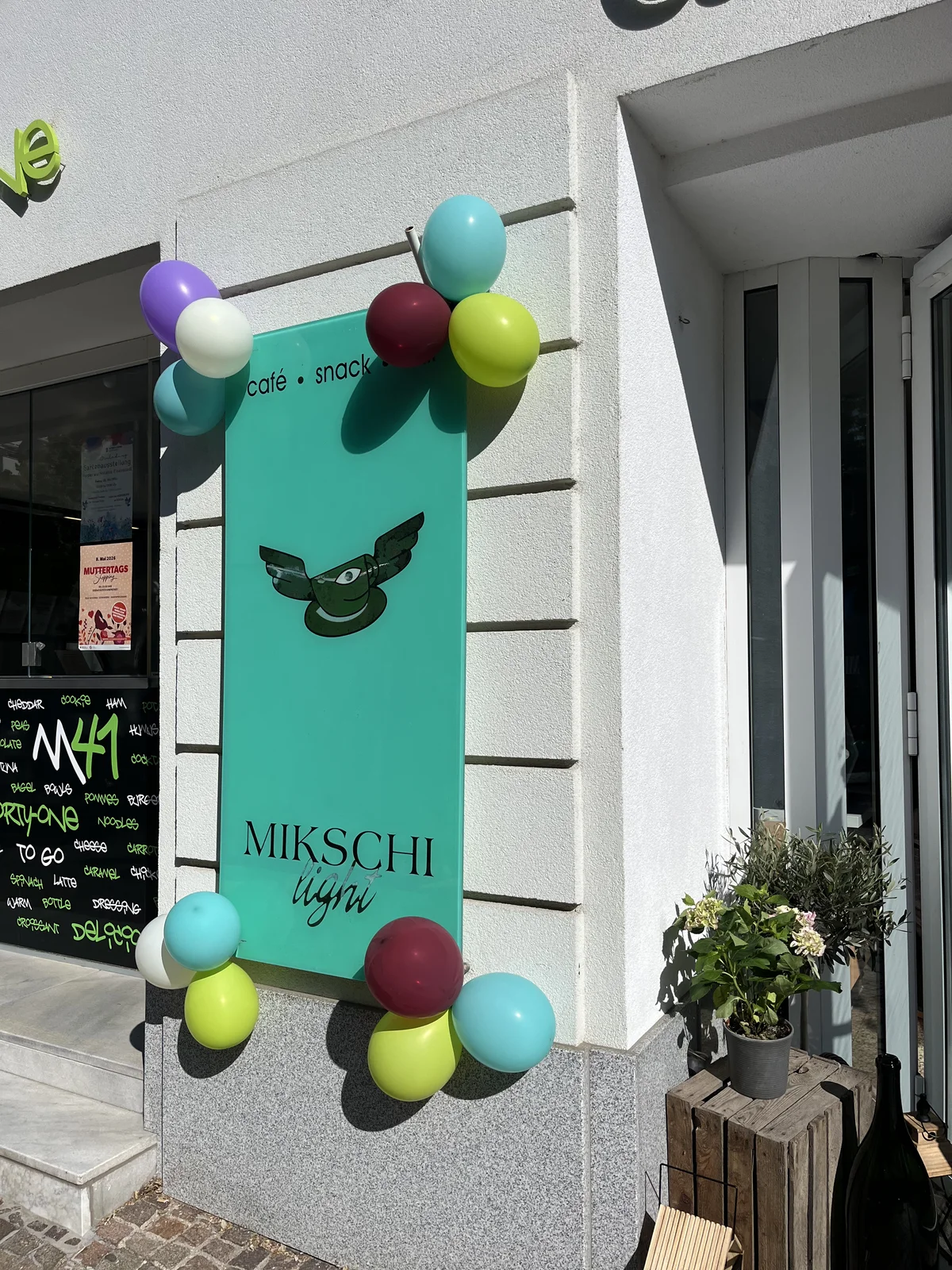Türkises Mikschi-Schild mit bunten Luftballons neben dem Eingang